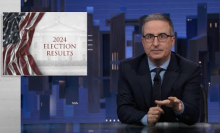 A man in a suit sits behind a talk show desk.